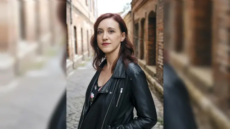 Eine junge Frau mit rötlichem Haar trägt eine schwarze Lederjacke vor einer Backsteinkulisse.
